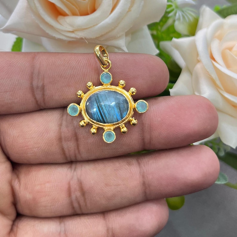 May include: A gold pendant with an oval, blue-grey stone at the center, surrounded by small, light blue stones and gold accents. The pendant is held up by a gold loop. The design is ornate and detailed, suitable for jewelry.