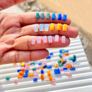 Puede incluir: Surtido de pequeños cabujones de ópalo rectangulares e iridiscentes en varios colores, incluidos azul, verde, amarillo y blanco. Los ópalos están dispuestos en filas en una mano y una superficie blanca.