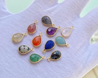 May include: A collection of teardrop-shaped gemstone pendants in various colors, including red, purple, blue, and green. Each pendant has a gold-colored bezel and a small loop for attaching to a chain or other jewelry component. The gemstones are displayed on a white surface.