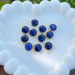 May include: A collection of round, faceted, deep blue gemstone pendants with gold-colored bezels and small loops for stringing. The pendants are arranged on a white, heart-shaped surface.