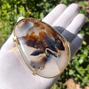 May include: A large, oval-shaped agate stone with brown, white, and black swirling patterns is set in a gold metal frame. The stone is smooth and polished.