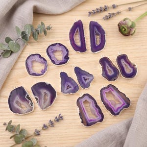 May include: A collection of polished, purple agate pendants with silver-colored edges. The pendants vary in shape and size, showcasing the natural patterns and colors of the stone. Some have a hollow center, while others are solid.