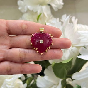 Op de afbeelding: Een hartvormige hanger met een dieprode edelsteen, met gesneden details en een kleine, ronde, heldere steen in het midden. De hanger is omlijst met goudkleurige accenten en een gouden balein. De hanger wordt in een hand gehouden.