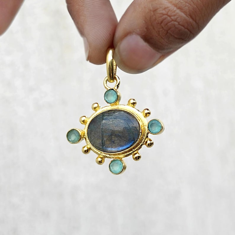 May include: Gold-tone pendant with an oval, iridescent, gray-blue stone at the center, surrounded by small, light blue stones and gold accents. The pendant is held up by a gold loop.
