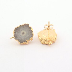 May include: A pair of gold-colored stud earrings. Each earring features a gray, circular stone with a lighter center, set in a gold-colored, textured frame. The earrings are displayed against a white background.