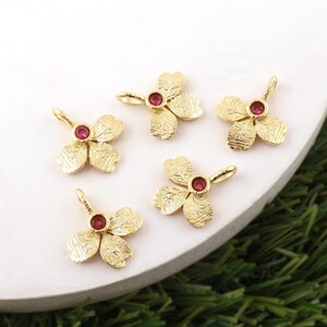 May include: Five gold-colored flower charms with a textured finish. Each charm features three petals and a small, round, red gemstone in the center. The charms are arranged on a white surface with green grass visible in the background.