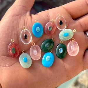 Può includere: Una collezione di ciondoli ovali in pietre preziose di vari colori, tra cui blu, verde, rosa e trasparente. Ogni ciondolo ha un piccolo anello dorato nella parte superiore. Alcuni ciondoli presentano una pietra a contrasto al centro.