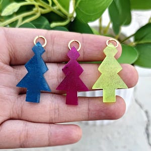 May include: Three Christmas tree-shaped pendants in blue, magenta, and yellow-green. Each pendant has a gold-colored loop at the top. The pendants are held in a hand, with a blurred background of green leaves and a white pot.