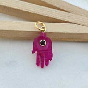 May include: A magenta-colored Hamsa hand charm with a gold-colored loop. The charm features a black circular stone in the center. The hand has carved details and is displayed on a light-colored surface.