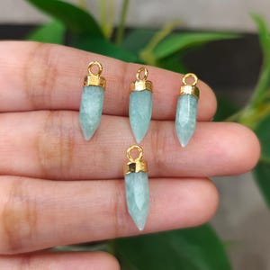May include: Four light blue gemstone pendants with gold-colored metal caps and loops. The pendants are faceted and have a pointed shape. The background is blurred, with green foliage visible.