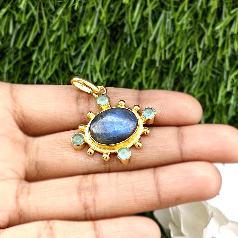 May include: Gold-colored pendant with a central oval, iridescent blue stone, surrounded by smaller, light blue stones. The pendant has a sunburst design with a loop at the top for a chain. The pendant is held in a hand.