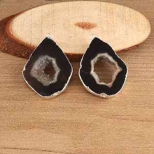 Op de afbeelding: Twee zwart-witte geode hangers met zilverkleurige rand. De hangers hebben een druppelvorm met een hol midden. De hangers worden gepresenteerd op een houten oppervlak.