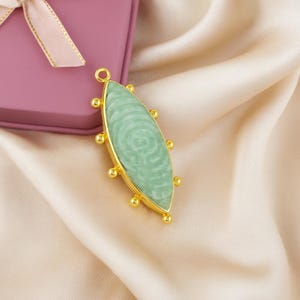Puede incluir: Colgante dorado con una piedra ovalada verde claro. La piedra tiene un patrón arremolinado y está enmarcada por un bisel dorado con pequeños detalles redondos. Tiene un lazo en la parte superior para una cadena.