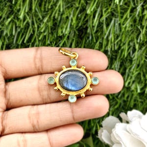 May include: A gold-colored pendant with a central oval-shaped blue gemstone, surrounded by smaller green gemstones. The pendant has a sunburst design and a loop for a chain. The background is a blurred green.