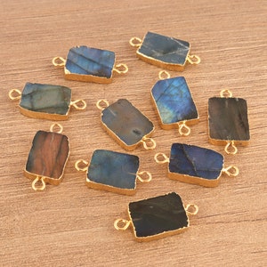 May include: A collection of ten rectangular labradorite gemstone connectors, each featuring a gold-toned metal edge and two small circular loops on opposite sides. The stones display varied shades of blue, gray, brown, and black with natural veining.
