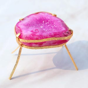 May include: A pink agate geode with a gold metal stand. The geode is a natural stone with a crystal formation inside. The stand has three legs and is made of gold metal.