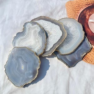 May include: Set of five agate coasters with gold trim. The coasters are various shades of gray and white with a natural agate pattern.