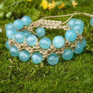 Op de afbeelding: Een handgemaakte armband met lichtblauwe, ronde kralen geregen op een beige macramé band. De armband ligt op een bedje van groen mos, met gele bloemen op de achtergrond. De kralen zijn ongeveer 1 cm in diameter.