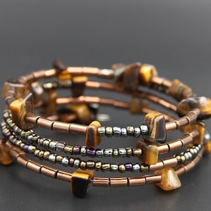 Könnte beinhalten: Ein Spiralarmband mit Tigeraugensteinen und kupferfarbenen Perlen. Das Armband hat mehrere Stränge mit kleinen, metallischen Perlen und größeren, rechteckigen Tigeraugensteinen. Das Design ist erdig und natürlich.