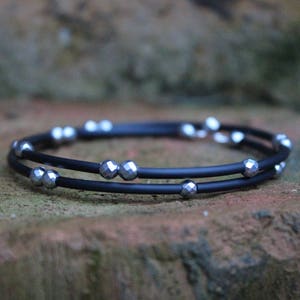 May include: A black wrap bracelet with two strands. The bracelet is adorned with multiple silver-colored, faceted beads. The bracelet is resting on a textured, brown surface.