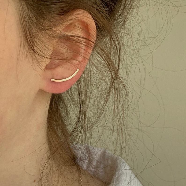 Puede incluir: Un pendiente de aro dorado con un dise&ntilde;o de barra curva. El pendiente se lleva en la parte superior de la oreja.