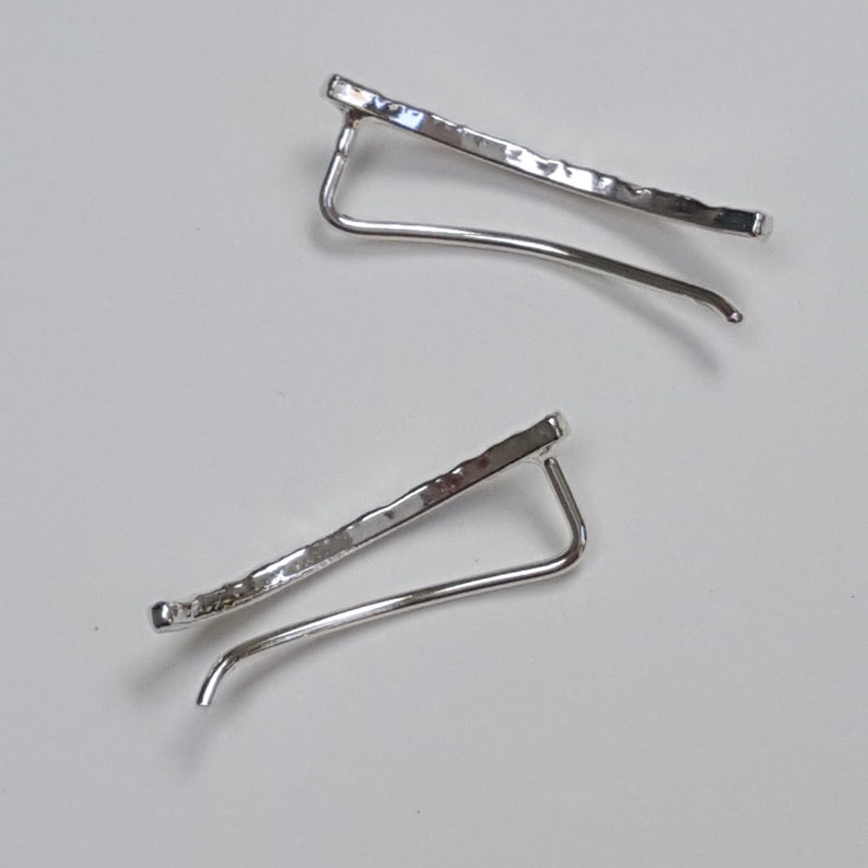 Puede incluir: Un par de pendientes de aro de plata con un acabado texturizado martillado. Los pendientes son curvos y tienen un dise&ntilde;o simple y minimalista.