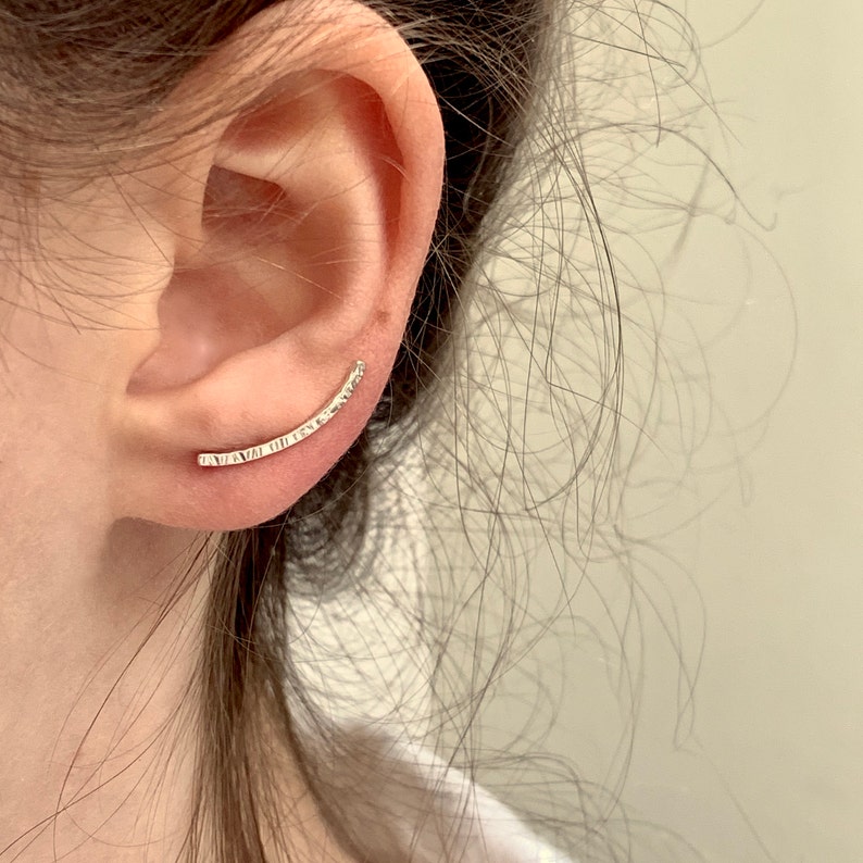 Puede incluir: Un pendiente de oreja de plata con una superficie texturizada. El pendiente es curvo y se coloca en la parte superior de la oreja.