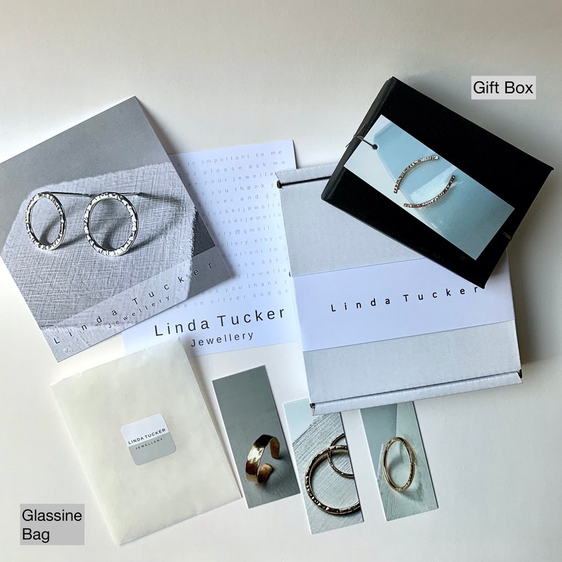 Puede incluir: Una caja de regalo blanca con el texto "Linda Tucker Jewellery" en la parte superior. La caja est&aacute; abierta y contiene una peque&ntilde;a bolsa blanca, una tarjeta con un brazalete de oro y dos tarjetas con anillos de plata. La caja est&aacute; sobre una superficie blanca. Hay una tarjeta blanca con el texto "Linda Tucker Jewellery" y una peque&ntilde;a bolsa blanca con el texto "Linda Tucker Jewellery" en ella. Tambi&eacute;n hay una tarjeta blanca con el texto "Gift Box" y una tarjeta blanca con el texto "Glassine Bag".