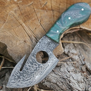 May include: A handmade knife with a green and white patterned handle and a Damascus steel blade. The blade has a hole in the center and a small hook on the side.