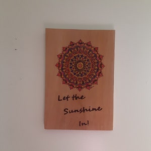 May include: A wooden sign with a mandala design in the centre, featuring a vibrant array of colours including red, orange, yellow, and purple. The sign also has the text "Let the Sunshine In!" written in black script.