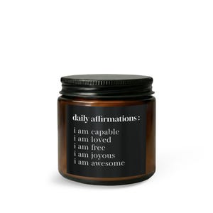 May include: A brown glass candle jar with a black screw-top lid and a black rectangular label. The label features the white text: daily affirmations: i am capable, i am loved, i am free, i am joyous, i am awesome, printed in a serif font.