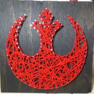 May include: Red string art of the Rebel Alliance symbol from Star Wars. The symbol is made of red string wrapped around nails hammered into a wooden board.