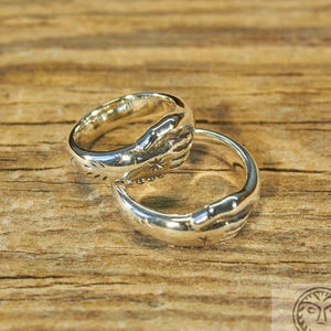 May include: Two silver rings with a design of clasped hands. The rings are on a wooden surface.