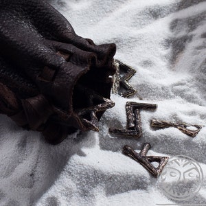 May include: A brown leather pouch with a drawstring closure, spilling out silver metal runes. The pouch is sitting on a white surface.
