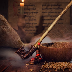 Peut inclure: Une pipe à fumer en bois avec un long manche fin et une coupe sculptée. La pipe est décorée de perles rouges et repose sur une surface en bois avec un bol et des grains. La pipe se trouve dans une pièce faiblement éclairée avec une bougie en arrière-plan.