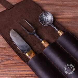 Puede incluir: Una funda de cuero marrón con tres ranuras contiene un cuchillo, un tenedor y una cuchara. Los utensilios tienen mangos de madera y hojas de metal.