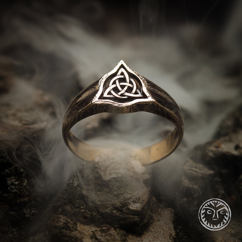 K&ouml;nnte beinhalten: Ein goldener Ring mit einem dreieckigen Design, das einen keltischen Knoten zeigt. Der Ring ist vor einem dunklen Hintergrund mit Rauch platziert.
