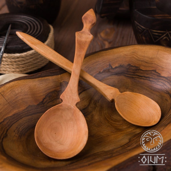 May include: Two wooden spoons with ornate handles, one with a rounded bowl and the other with a more pointed bowl, are crossed over each other in a wooden bowl.