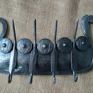 Pode incluir: Porta-chaves de metal preto em forma de navio viking com cinco ganchos. O navio tem uma cabeça de dragão na proa e escudos nas laterais.