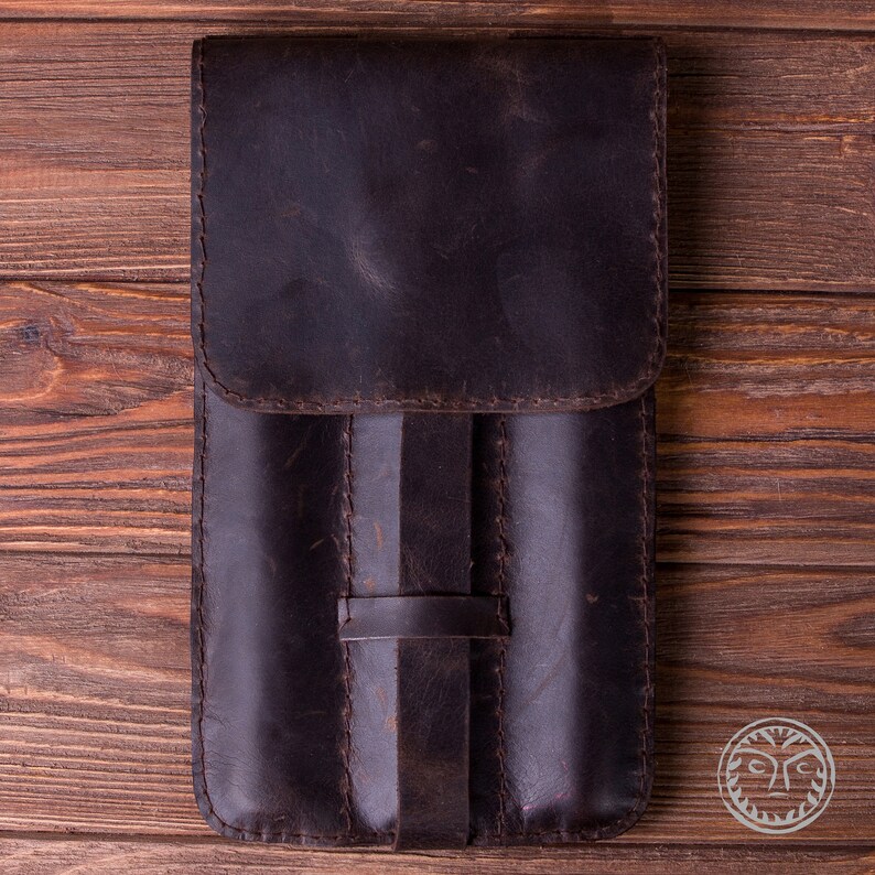 May include: A brown leather case with three slots for holding pens or pencils. The case has a flap that closes with a strap.