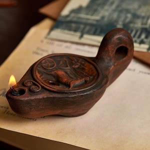 May include: A terracotta-colored oil lamp with a detailed relief design, lit with a small flame. The lamp features a circular reservoir with an embossed figure and decorative swirls. The background includes aged paper and a vintage postcard.