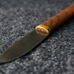 Puede incluir: Un cuchillo peque&ntilde;o con mango negro, una virola de lat&oacute;n y un mango de madera. La hoja es curva y tiene una punta afilada.