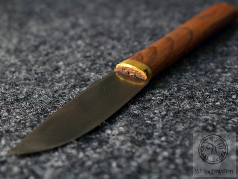 Puede incluir: Un cuchillo peque&ntilde;o con mango negro, una virola de lat&oacute;n y un mango de madera. La hoja es curva y tiene una punta afilada.
