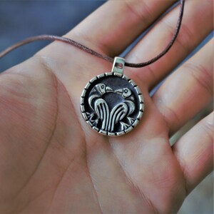 May include: A silver pendant necklace with a circular design featuring two birds facing each other with their beaks touching. The pendant is on a brown cord.