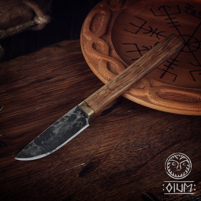 Puede incluir: Un peque&ntilde;o cuchillo con mango negro, mango de madera y un acento de lat&oacute;n. El cuchillo est&aacute; sobre una superficie de madera con un plato de madera tallado en el fondo.