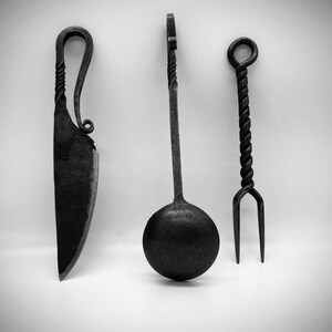 May include: A set of three black metal kitchen utensils: a knife, a spoon, and a fork. The utensils have a twisted design and are handcrafted.