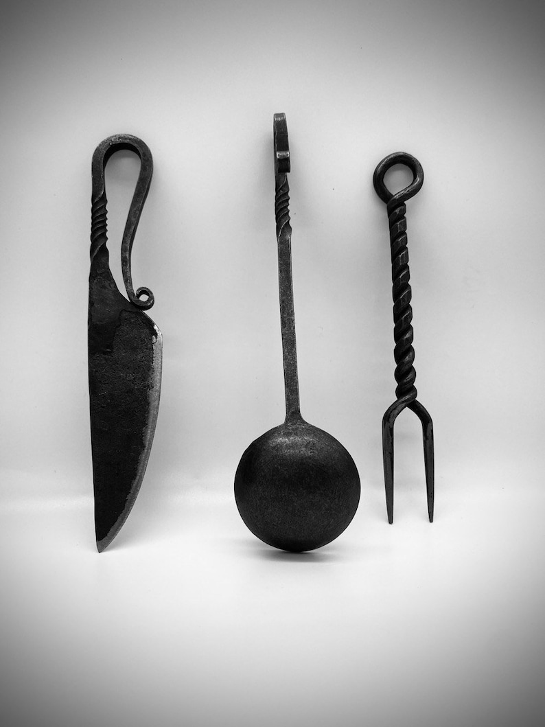 May include: A set of three black metal kitchen utensils: a knife, a spoon, and a fork. The utensils have a twisted design and are handcrafted.