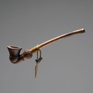 Puede incluir: Una pipa de cerámica marrón con un tallo largo y delgado de madera. La pipa tiene un diseño decorativo y está atada con un cordón marrón con cuentas negras.