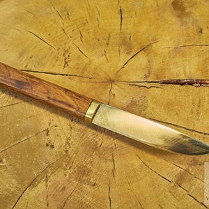 Puede incluir: Un cuchillo con mango de madera y hoja de lat&oacute;n, que se encuentra sobre una superficie de madera.