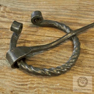 May include: A twisted metal Viking brooch with a decorative loop at the top. The brooch is made of dark metal and has a rustic finish.